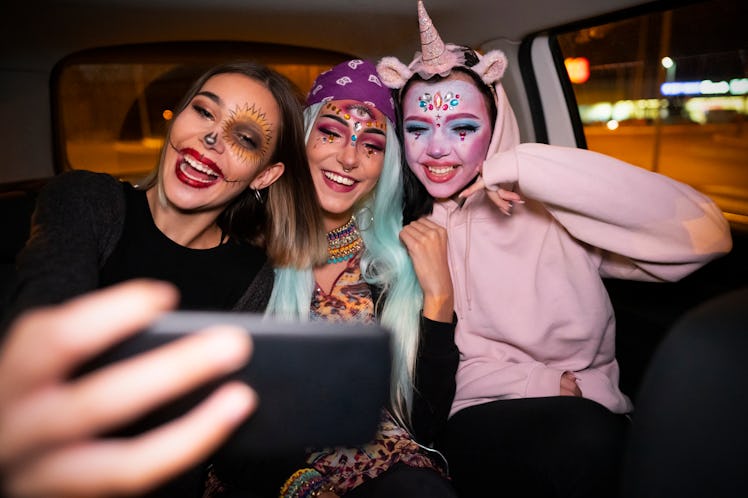 Three young women dressed in Halloween costumes take a selfie while sitting in the backseat of a car...