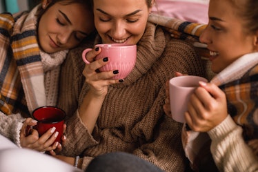 Three women wearing cozy oversized sweaters hold up their coffee mugs and cuddle.