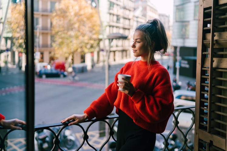 A woman holding a coffee mug on her balcony is dressed in a red turtleneck sweater and black jeans.
