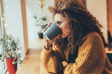 A woman in a gold sweater sips a black mug in a bright coffee shop while looking out the window.
