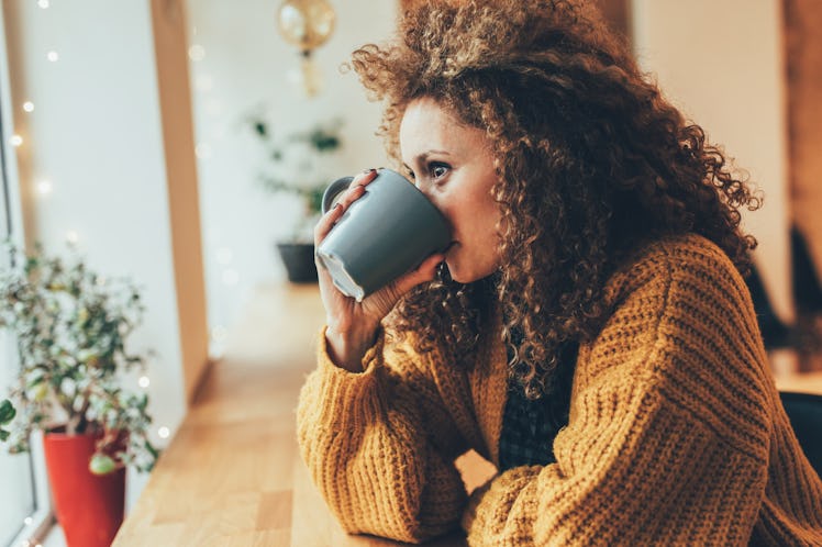 A woman in a gold sweater sips a black mug in a bright coffee shop while looking out the window.