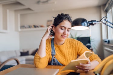 A young woman reads a book while sitting next to her laptop and wearing a yellow sweater.