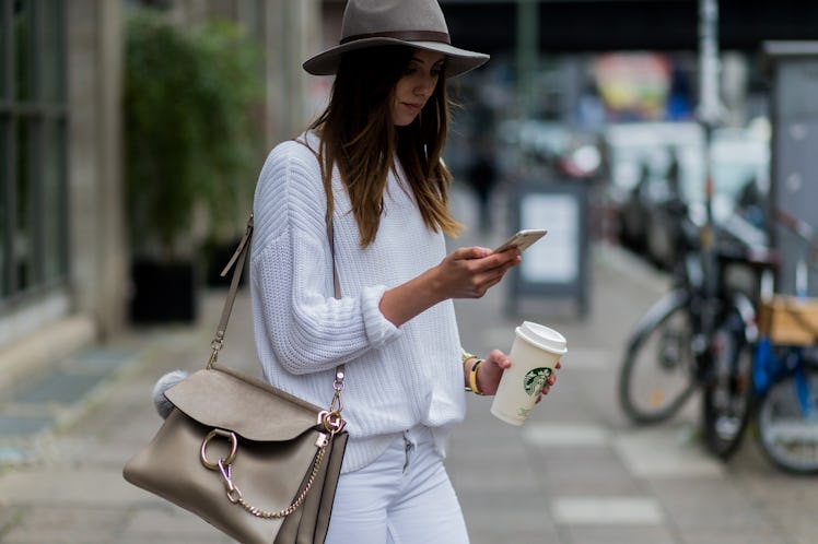 A woman in a felt hat, white sweater, and white jeans holds her Starbucks cup while texting on a cit...