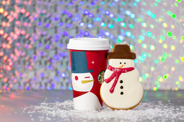 A holiday Starbucks cup is placed on fake snow with a snowman cookie next to it.