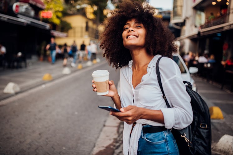 A happy, trendy woman dressed in a white button-down, blue jeans, and black leather backpack looks d...