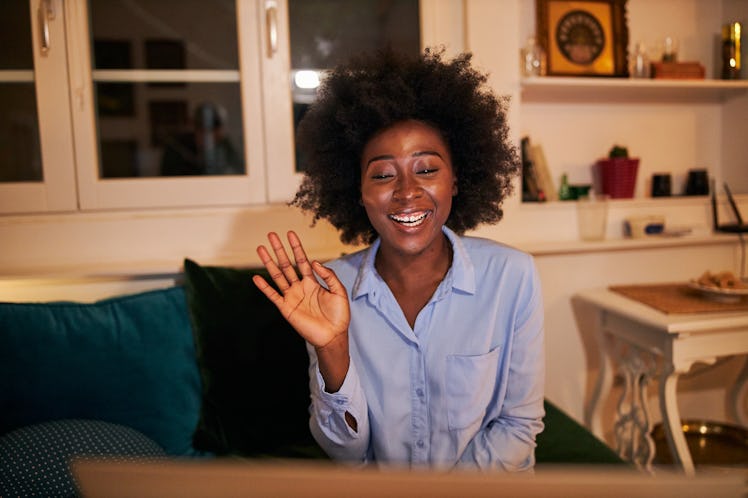 A young Black woman waves at her laptop while video chatting with her long-distance best friend.