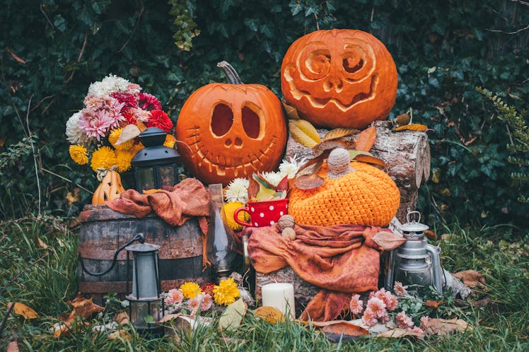 Two jack-o-lanterns are placed on a rustic setup with flowers, lanterns, fall leaves, and a blanket.