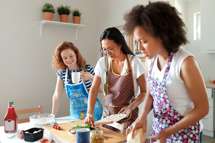 Three women in aprons make pizza in a bright kitchen.