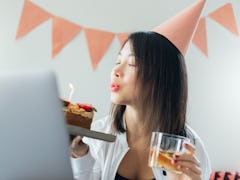 A young Asian woman blows out a candle on her birthday cake while video chatting with her friends.