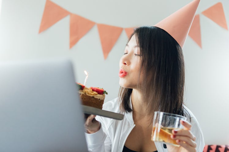 A young Asian woman blows out a candle on her birthday cake while video chatting with her friends.