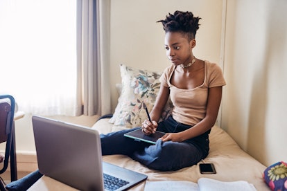A young Black woman draws on a tablet while looking at her laptop and sitting on her bed.