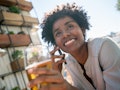 A young woman sits outside her apartment building and holds a beer while talking on the phone.