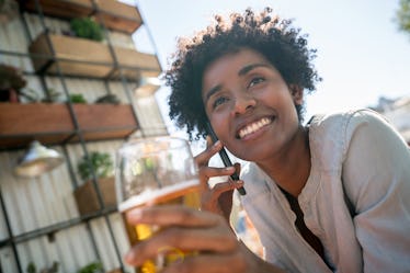 A young woman sits outside her apartment building and holds a beer while talking on the phone.