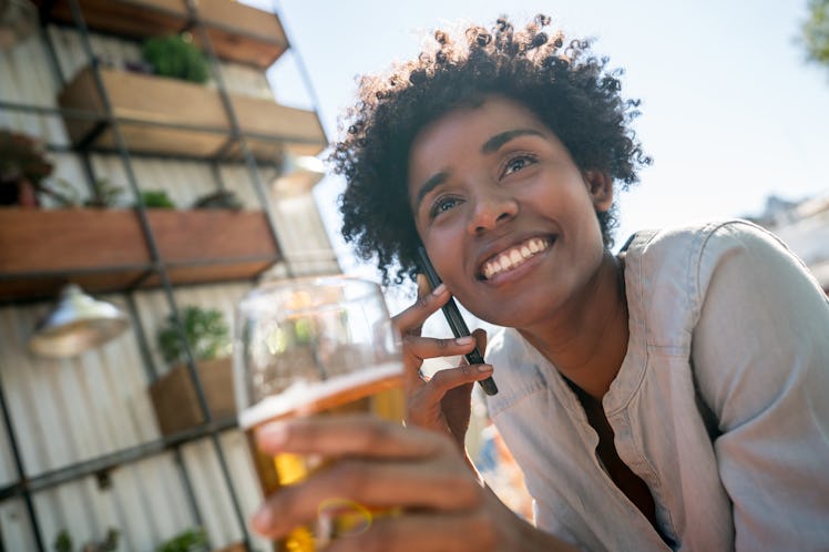 A young woman sits outside her apartment building and holds a beer while talking on the phone.
