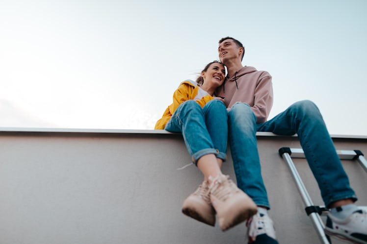 A young couple laughs while sitting on a rooftop together at sunset during the fall.