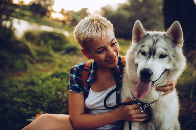 A young woman with short hair sits with her husky while on a fall hike at sunset.