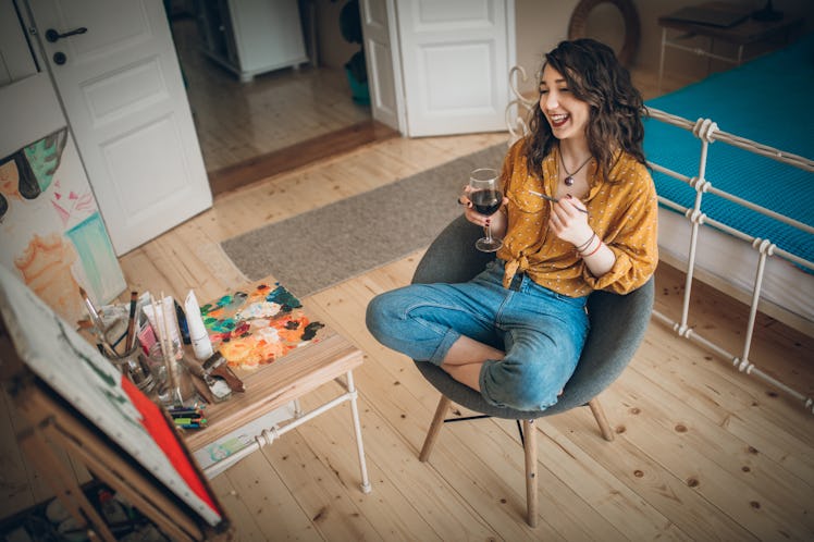 A happy woman paints a canvas with a glass of wine in hand, while sitting in her bedroom. 