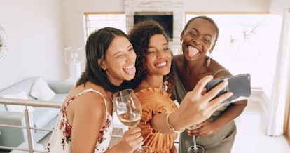 Three best friends smile for a selfie, while holding wine in their hands. 
