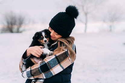 A happy woman bundled up for winter hugs her puppy in the snow.