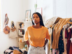 A young Black woman stands in front of a rack of trendy clothes in her home.