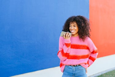 A young Black woman stands in front of a blue and red wall while wearing a colorful sweater and hold...