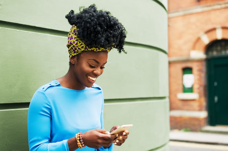 A happy woman wearing a blue long-sleeve shirt smiles while she texts on a sunny day.