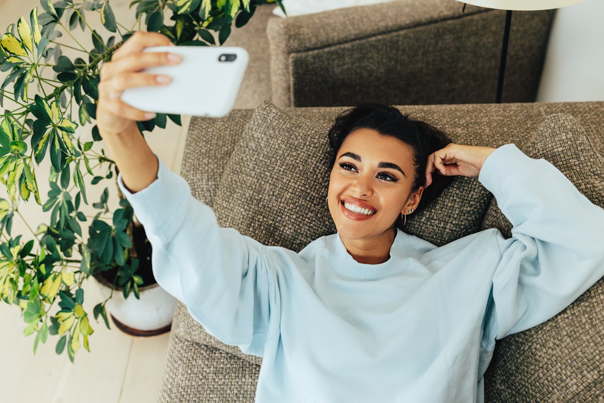 A beautiful woman wearing a light blue sweatshirt takes a selfie on her couch in her bright living r...
