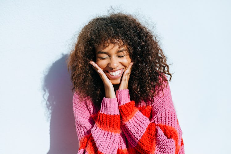 A young woman with curly hair smiles wide for a camera, while wearing a pink and red sweater.