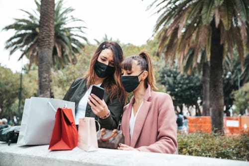 Two people wearing masks look at one of the people's phone outside near palms trees. Keep your masks...