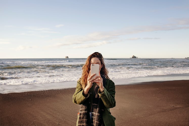 A young woman stands in front of an ocean in the winter and holds up her phone.