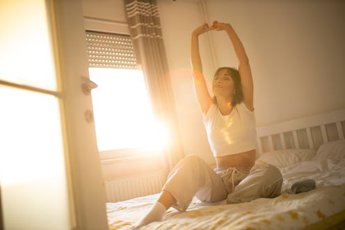 A person sits up in bed and stretches with light streaming through the window. Waking up can be toug...