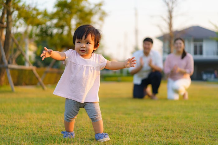 A toddler, bathed in sunlight, walks across a lawn, her parents visible in the background.