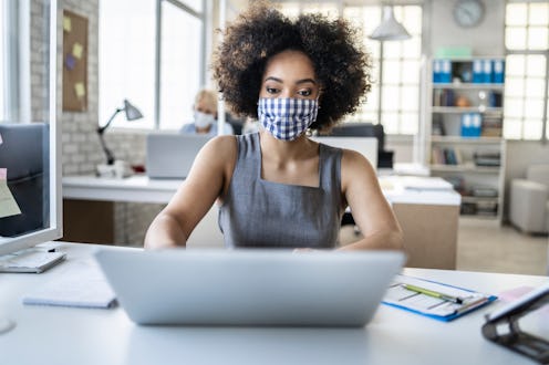 A woman wearing a gingham mask works at a laptop. Can your job force you to get vaccinated for covid...