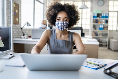 A woman wearing a gingham mask works at a laptop. Can your job force you to get vaccinated for covid...