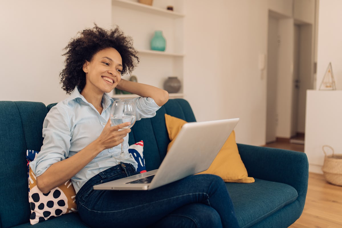A happy woman enjoys wine and a virtual event on her blue couch.