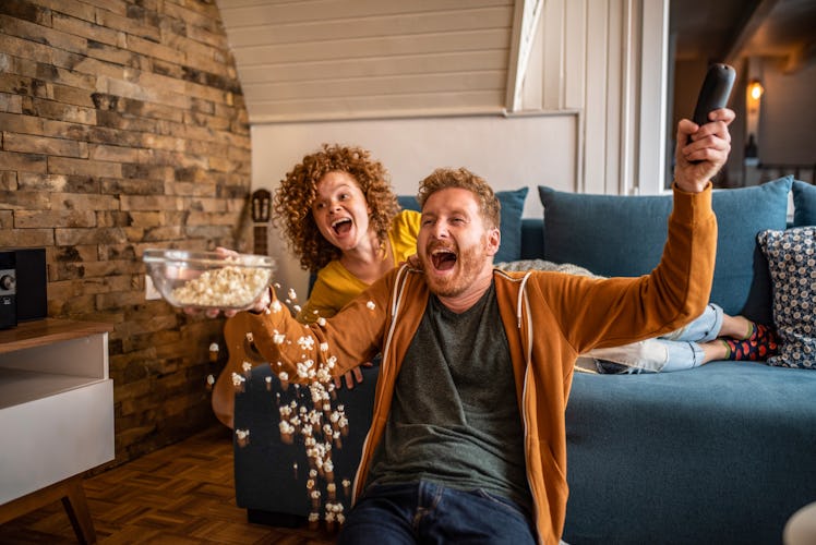 A young couple cheers in their home while eating popcorn and watching the Super Bowl.