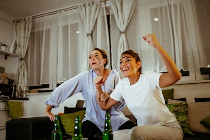 Two women cheer while watching the Super Bowl on their couch and doing face masks.