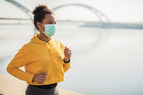 A woman in a yellow track jacket jogs with a mask on. How covid vaccines will affect your mental hea...