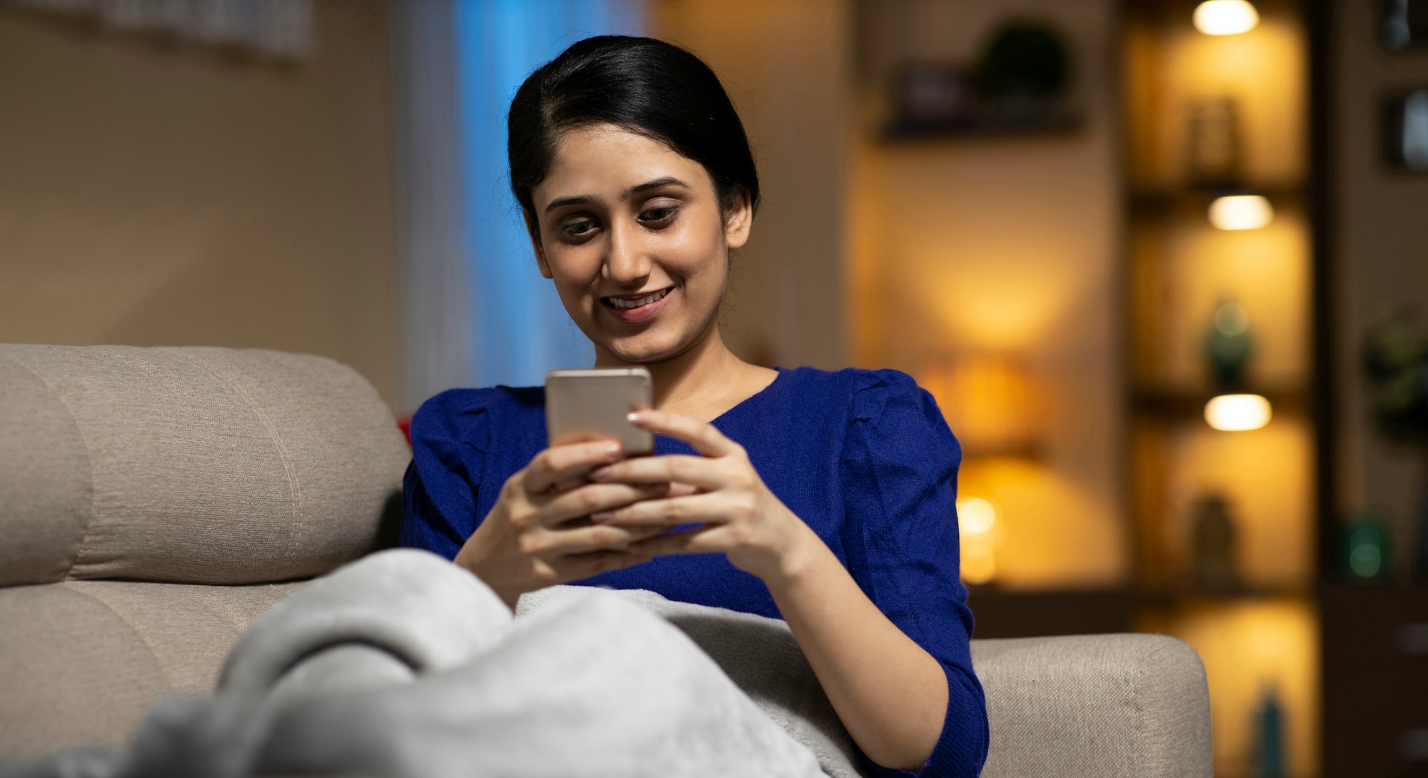 A black-haired lady smiling on a couch under a blanket and typing text messages on her mobile phone