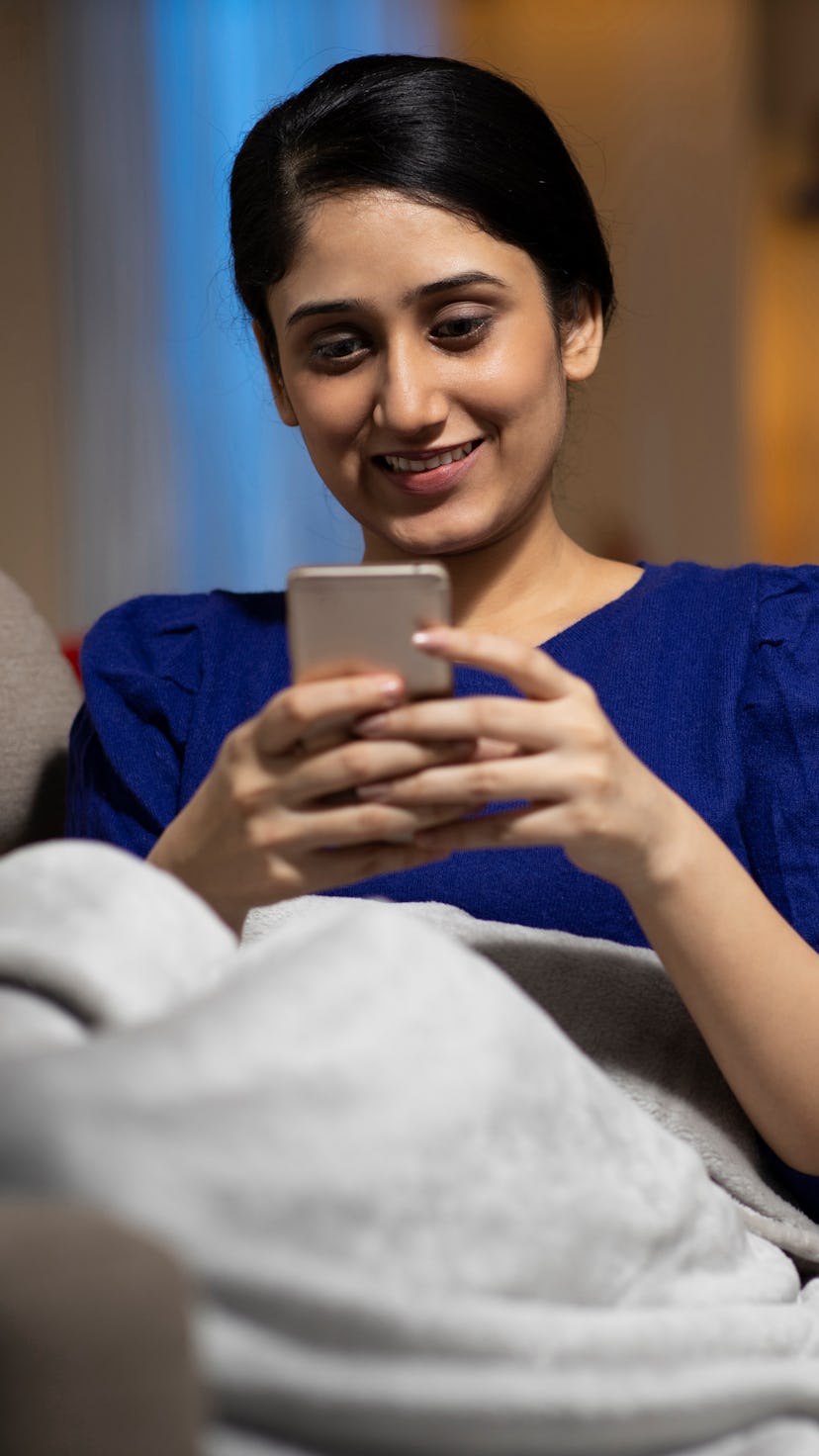 A black-haired lady smiling on a couch under a blanket and typing text messages on her mobile phone