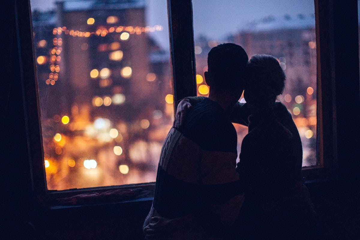 A young couple cuddles next to a window in their apartment on Valentine's Day and looks out at the city lights.
