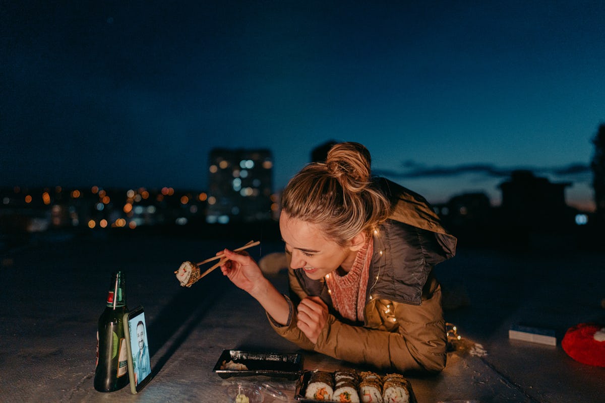 A happy woman smiles while enjoying sushi on a rooftop, on a virtual Valentine's Day date with her partner.