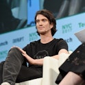 WeWork CEO Adam Neumann is seen at a panel. He is wearing a microphone and sitting on a couch.
