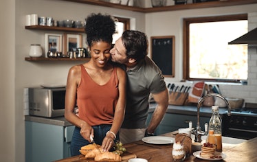 A happy woman cuts a croissant in a bright kitchen while her husband gives her a kiss.