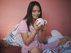 A happy woman in her striped pajamas looks into her coffee mug on her bed.