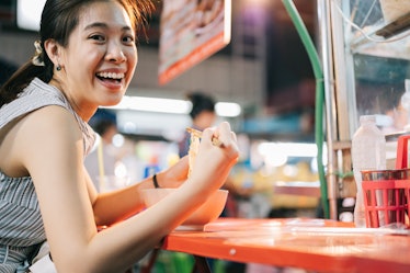 A happy woman eats noodles from a vendor while on vacation. 