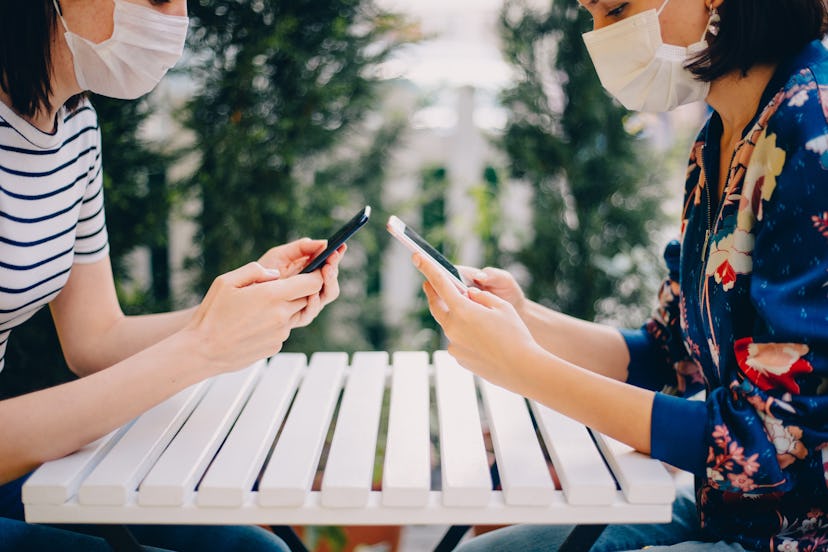 Two friends wear masks outside while on their phones. Doctors explain  Why Some People Don’t Get COV...