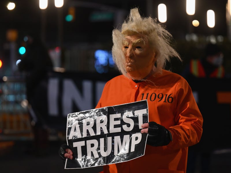 A man in a Trump mask holding a sign that says "arrest Trump."