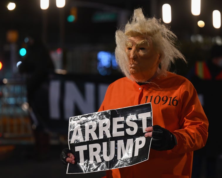 A man in a Trump mask holding a sign that says "arrest Trump."
