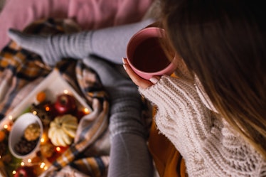 A woman wearing leggings and a cozy sweater sips a cup of tea on her bed while enjoying afternoon te...
